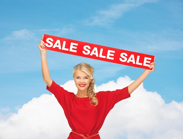 sale, shopping, christmas, holidays and people concept - smiling woman in red dress with red sale sign over blue sky and white cloud background