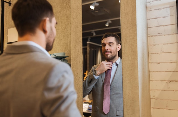 sale, shopping, fashion, style and people concept - happy young man choosing and trying tie on and looking to mirror in mall or clothing store