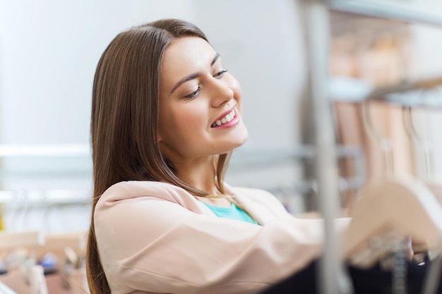 sale, shopping, fashion, style and people concept - happy young woman choosing clothes in mall or clothing store