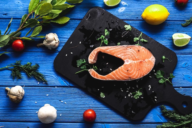 Salmon steak on a black cutting board and a blue wooden background, contrast, next to citrus vegetables and herbs