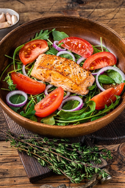 Salmon steak salad with green leaves arugula, avocado and tomato in a wooden plate