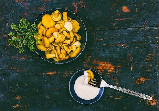 Salted milk mushrooms with green sprigs of dill in black bowl and mushroom on fork and sour cream in saucer on black wooden table with copy space