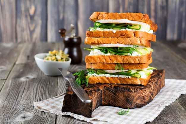 Sandwich for breakfast stuffed with slices of cucumber, curd cheese, ham and dill on old wooden background. Selective focus.