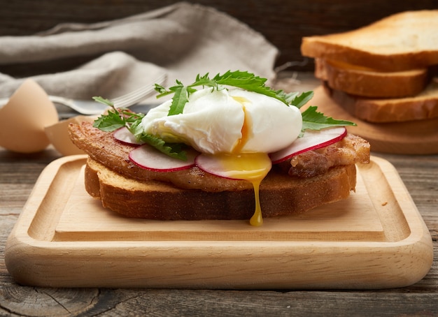 Sandwich on toasted white slice of bread with poached eggs, green leaves of arugula and radish