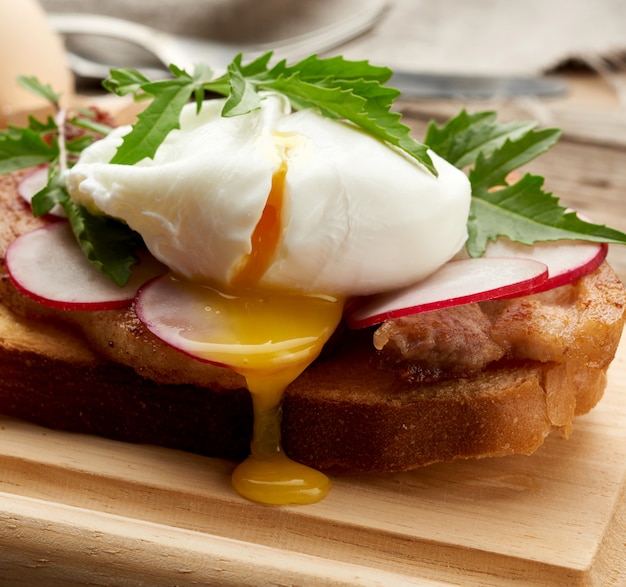 Sandwich on toasted white slice of bread with poached eggs, green leaves of arugula and radish