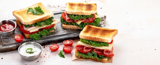 Sandwich with cheese ham tomato and fresh salad made of whole wheat bread with a crispy crust on a wooden board with sauces on a white background top view panorama with copy space