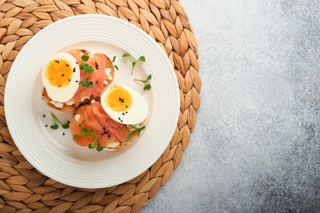 Sandwich with delicious toppings on top smoked salmon eggs herbs and microgreens radish black sesame seeds over white plate on concrete background Healthy open sandwich superfood Top view