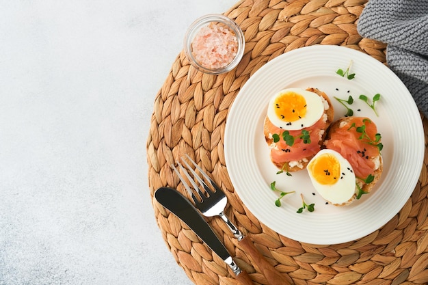 Sandwich with delicious toppings on top smoked salmon eggs herbs and microgreens radish black sesame seeds over white plate on concrete background Healthy open sandwich superfood Top view