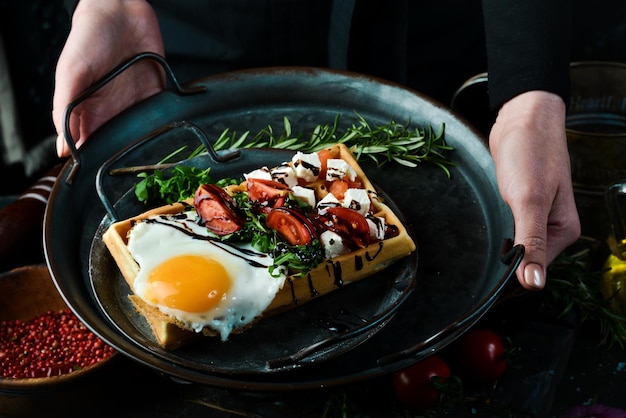 Sandwich with egg cheese salted fish and arugula on a plate in the hands of a chef on a rustic wooden background Belgian waffles On a black background