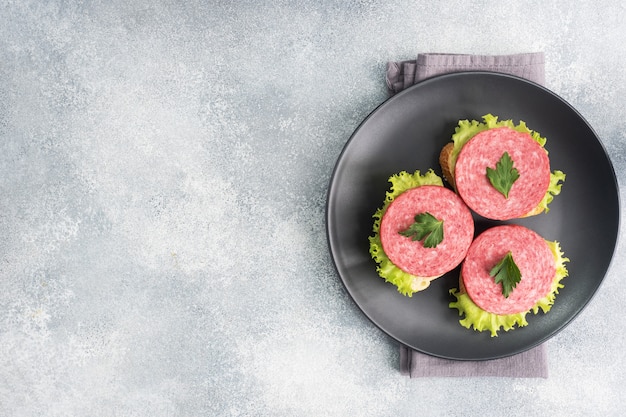 Sandwiches with lettuce leaves and sliced salami sausage on black plate. Copy space top view