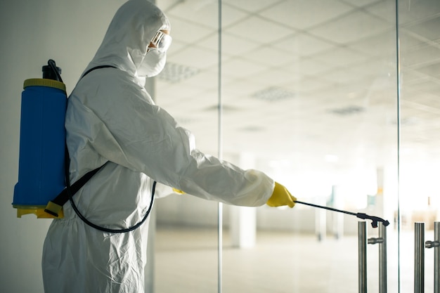 Sanitary worker sprays an empty business center with antiseptical liquid to prevent covid-19 spread. A man wearing disinfection suit cleaning up the shopping mall. Nobody, health, isolated concept.