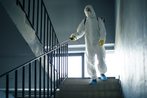 Sanitizing worker cleaning up the staircase at the shopping mall with an antiseptic to prevent covid-19 spread. A man in a disinfection suit sprays stairs. Healthcare, quarantine and hygiene concept.