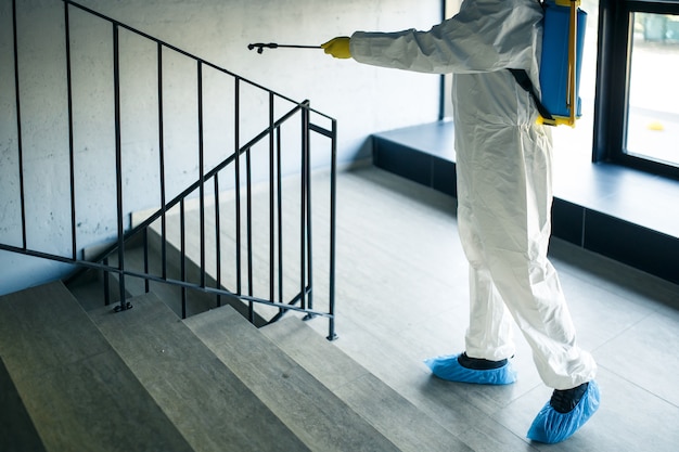 Sanitizing worker cleaning up the staircase at the shopping mall with an antiseptic to prevent covid-19 spread. A man in a disinfection suit sprays stairs. Healthcare, quarantine and hygiene concept.