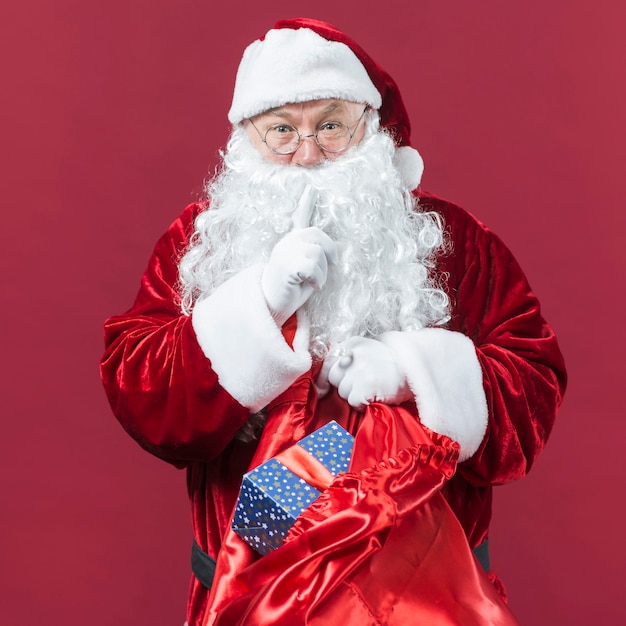 Santa Claus in glasses with sack of gifts showing secret gesture