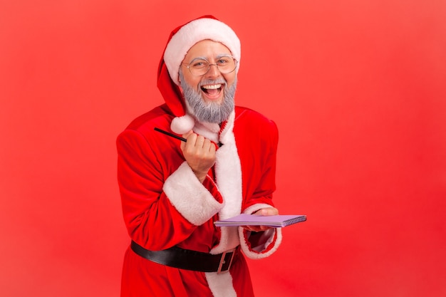 Santa claus holding paper notebook and pen in hands, laughing, looking at camera.