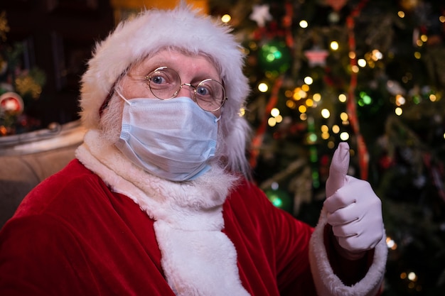 Santa Claus in medical mask. Portrait positive Santa showing thumb up sitting near Christmas tree