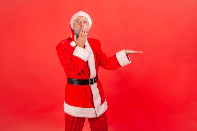 Santa claus presenting copy space on his palm, covering mouth with hand, astonished expression.