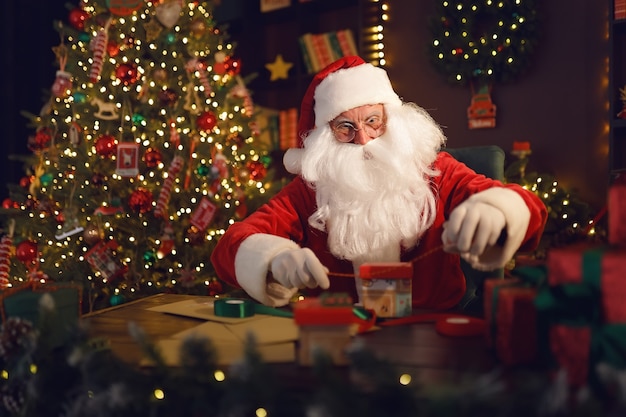 Santa claus or saint nicholas makes gifts while sits at the table at home near christmas tree