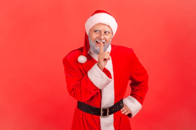 Photo santa claus shushing with silence gesture, finger on lips, smiling to camera, asking to keep secret.