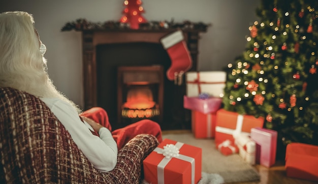 Santa claus sitting on the armchair near the christmas tree with presents at home