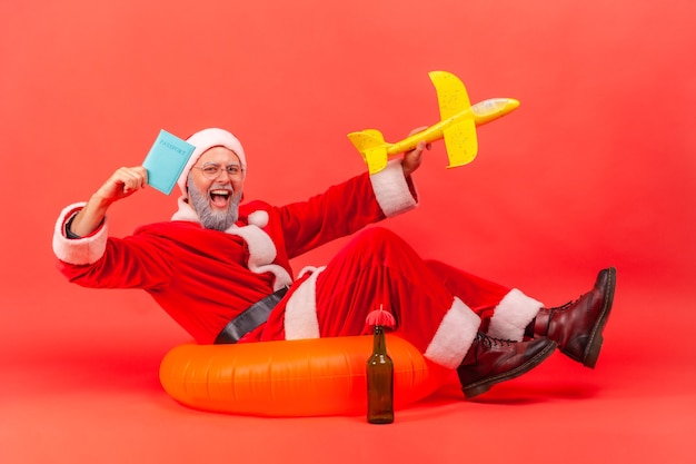 Santa claus sitting on rubber ring and holding paper plane and passport, enjoying winter traveling.