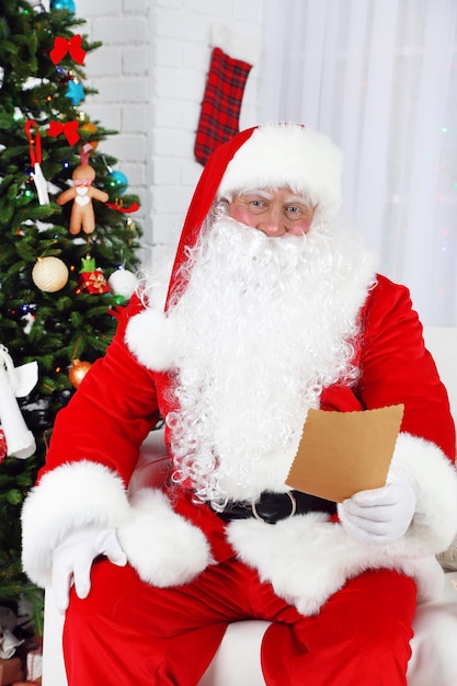 Santa Claus sitting on sofa with list of presents near Christmas tree