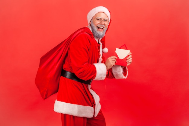 Santa claus standing with bag of presents and holding letter with Christmas congratulations.