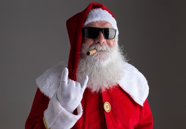 Photo santa claus wearing sunglasses showing the rocker hand sign and smoking a cigar on dark background