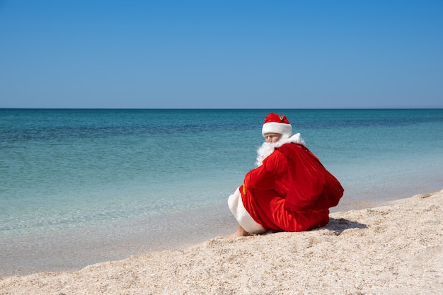 Santa Claus with a bag of gifts sitting on the sandy beach. Blue sky. Turquoise water. Copy space for your text - Image