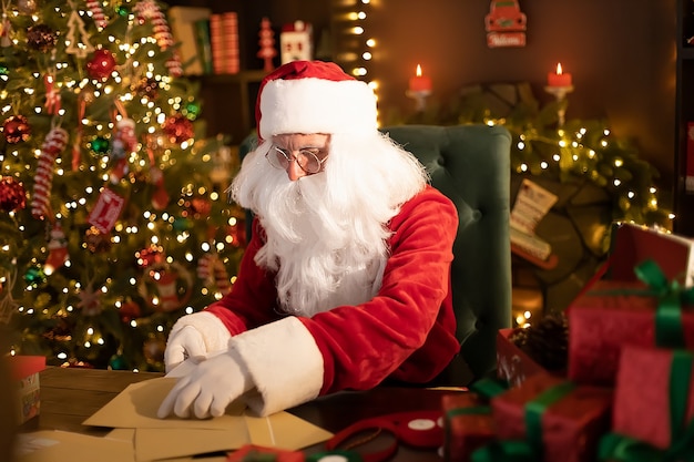 Santa clause is prepares gifts for children for xmas at his desk at home while opening wish lists