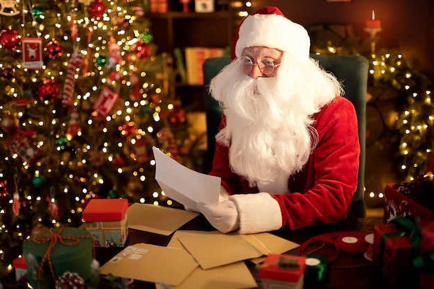 Santa clause is prepares gifts for children for xmas at his desk at home while reading wish lists