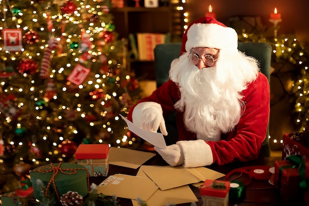 Santa clause is prepares gifts for children for xmas at his desk while reading wish lists at home