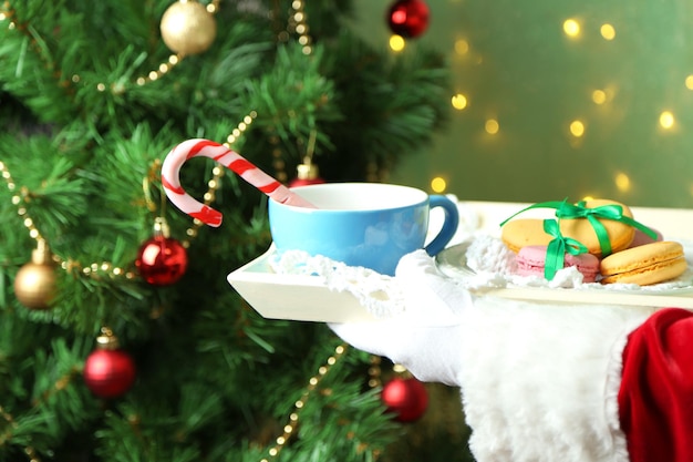 Santa holding mug and plate with cookies in his hand, on bright background