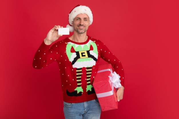 Santa man hold red gift box and credit card Studio portrait of guy wears santa hat and winter sweater Man with christmas hat over isolated red background