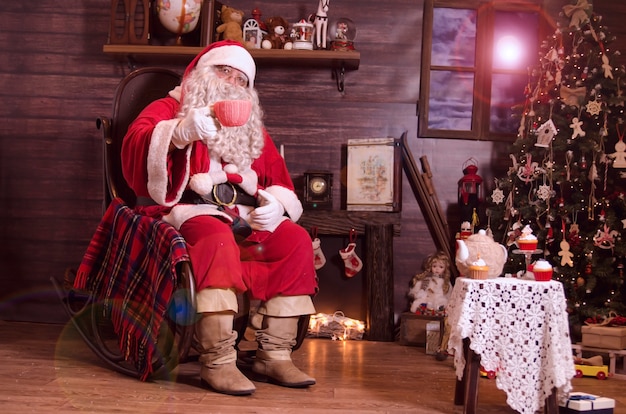 Photo santa sitting on rocking chair by the fireplace and cheers with big cup of milk in decorated room