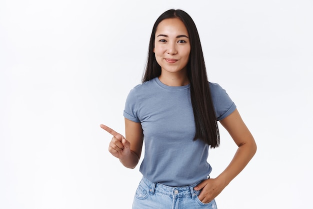 Sassy good-looking confident brunette asian female giving advice what product pick, showing best choice, pointing left, smirk satisfied give knowing look, hold hand in jeans pocket, white wall