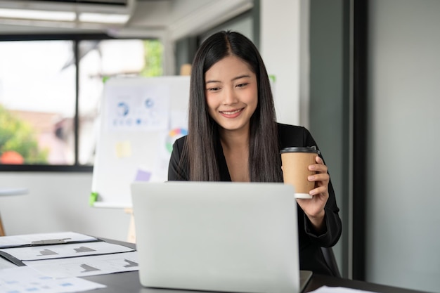 A satisfied Asian businesswoman is having a morning coffee while working on her laptop