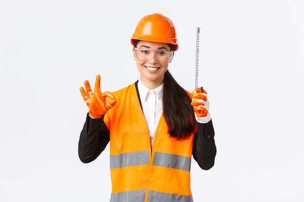 Satisfied asian female construction engineer, architect or inspector at enterprise showing okay gesture and tape measure, smiling pleased, give permission, approve measurements are valid