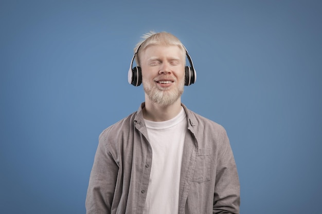 Satisfied calm albino guy in headphones enjoying favorite music with closed eyes standing over blue