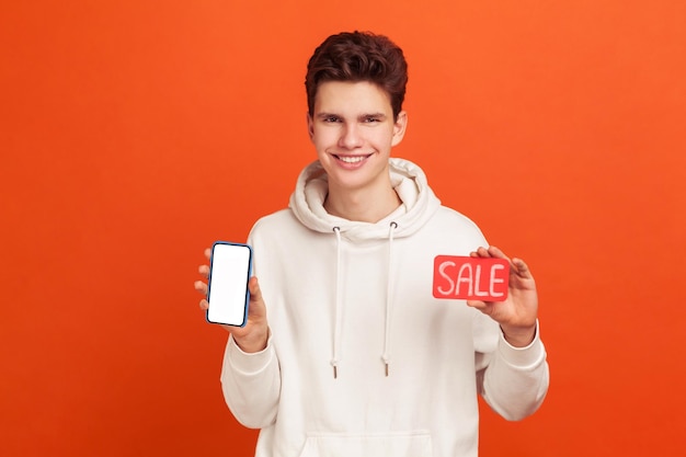 Satisfied cheerful teenager in casual style hoodie holding smartphone with blank display and sale card in hands, discount application. Indoor studio shot isolated on orange background