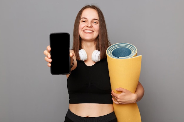 Satisfied delighted happy woman holding rolled fitness mat dressed in activewear isolated over gray background presenting mobile app for sporty workout empty display