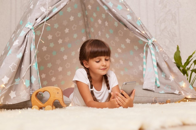 Satisfied delighted little girl with pigtails wearing white t shirt posing in wigwam at home and using phone watching cartoons or having video call expressing positive emotions