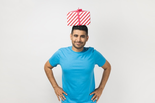 Satisfied delighted unshaven man standing with hands on hips holding present box on his head