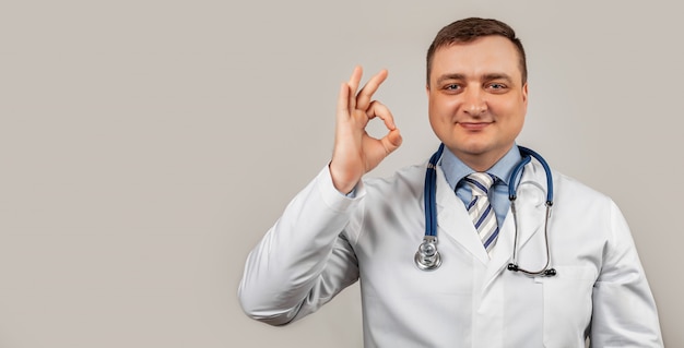 Satisfied doctor man in a medical coat and with a stethoscope shows okay okay sign of approval smiles, white background