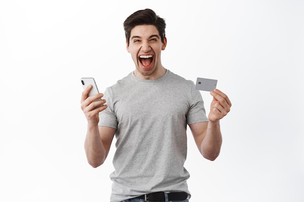 Satisfied guy celebrating, showing plastic credit card and mobile phone, screaming amazed, triumphing, winning money, standing over white background