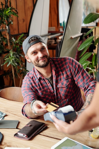 Satisfied handsome guest in ball cap paying for dinner with wireless card in modern cafe