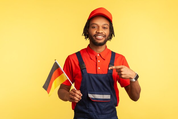 Satisfied handsome handyman in uniform with dreadlocks pointing finger at German flag in his hand and looking at camera with toothy smile. Indoor studio shot isolated on yellow background.