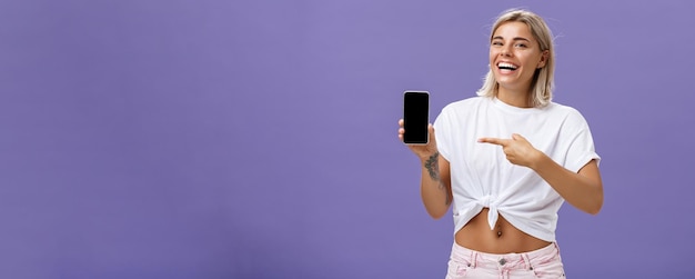 Satisfied happy beautiful tanned woman with blond hairstyle and tattoos holding smartphone showing