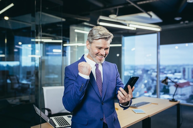 Satisfied and happy leader senior and experienced businessman looks at the phone smiles and rejoices in victory received good news