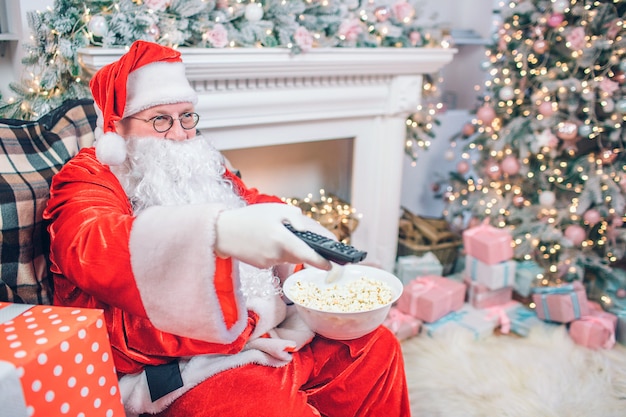 Satisfied and happy man in Santa Claus costume sits and uses remote control. He has bowl of popcorn in another hand. There are fireplace and Christmas tree behind man.